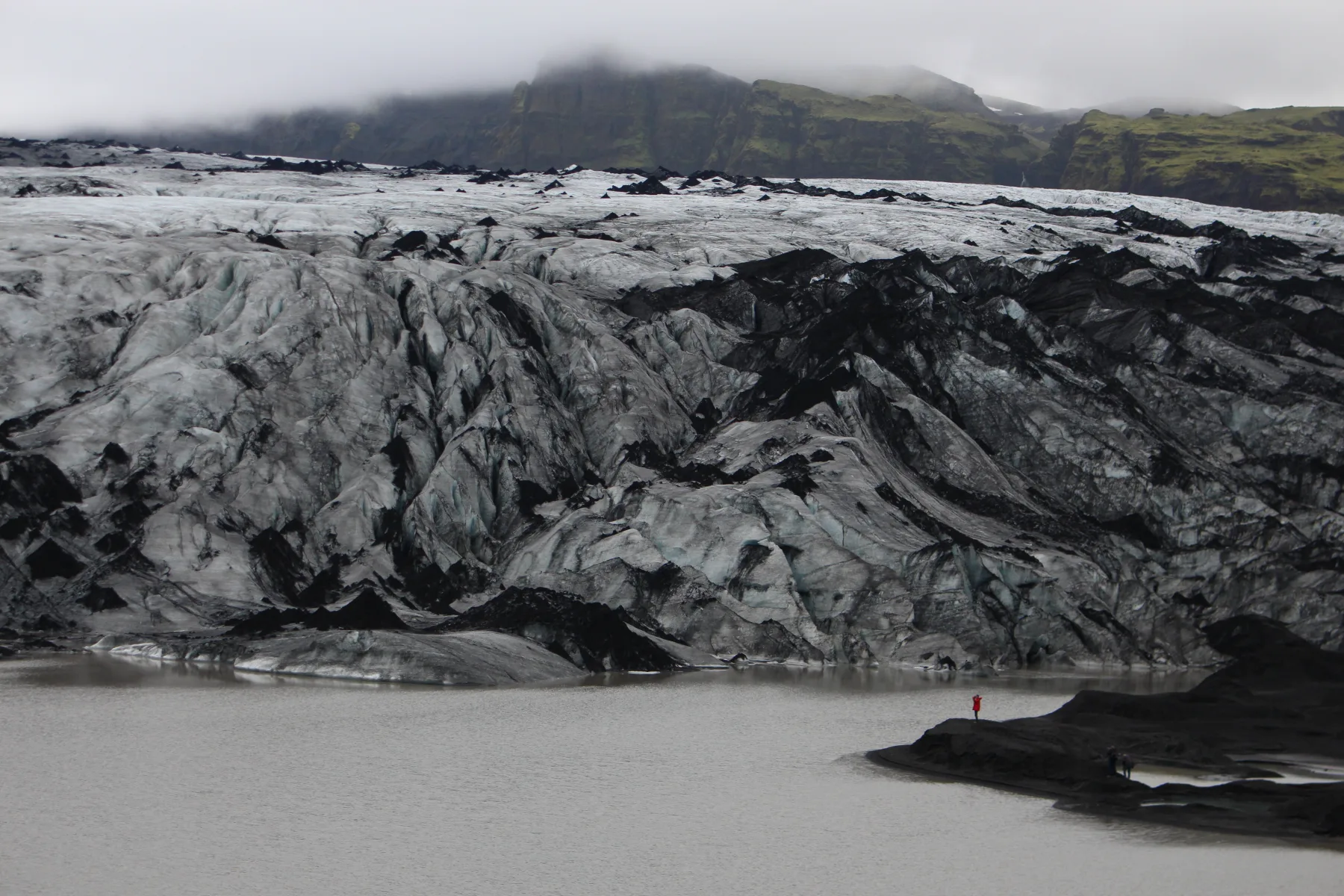 Glacier streaked with volcanic sediment beneath misty green cliffs